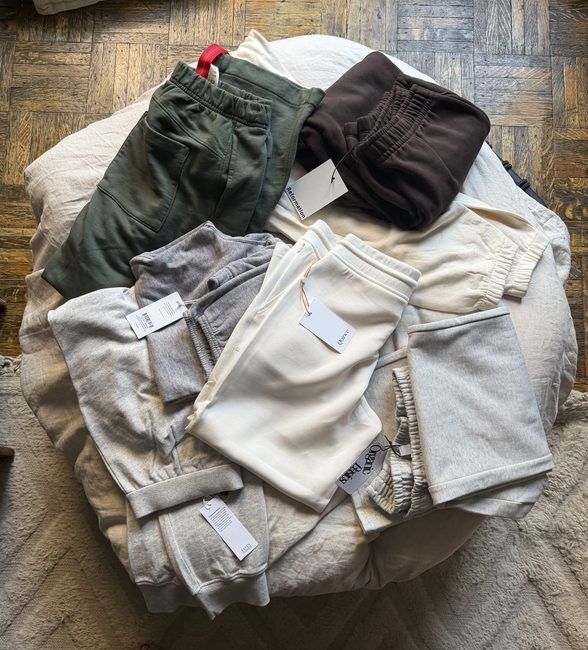 Assorted sweatpants and sweatshirts in neutral colors are arranged in a pile on a white cushion, each with brand tags attached, on a wooden floor.