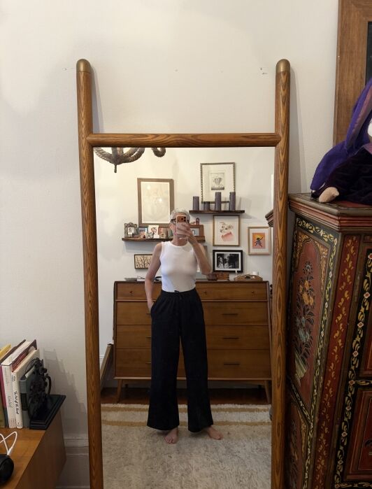 Person in a white sleeveless top and black pants takes a mirror selfie in a mid-century style room with framed art and wooden furniture.