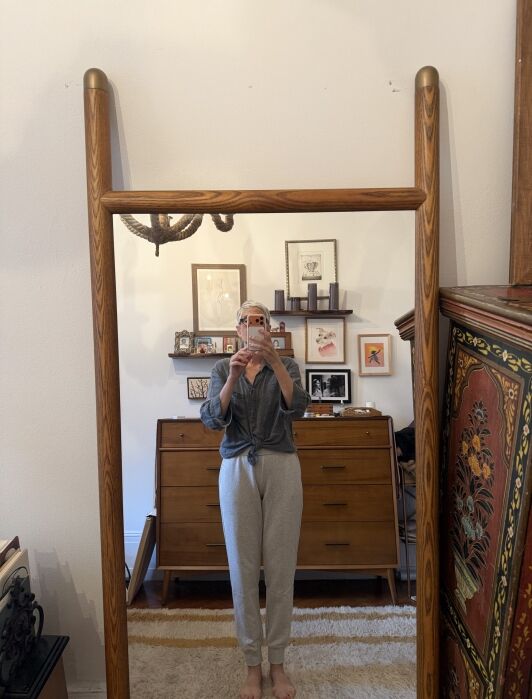 Person taking a mirror selfie in a cozy room with wooden furniture, framed art on the wall, and a patterned cabinet to the right.
