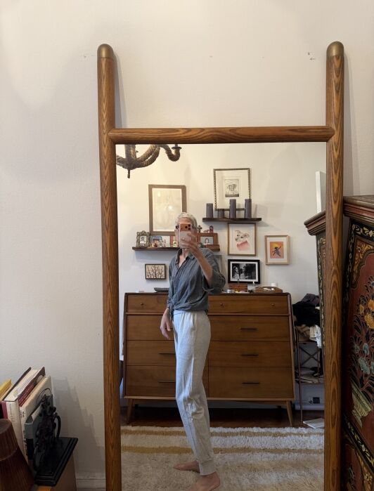Person taking a mirror selfie in a room with wooden furniture, art on the walls, and books on the floor; wearing a gray shirt and light gray pants.