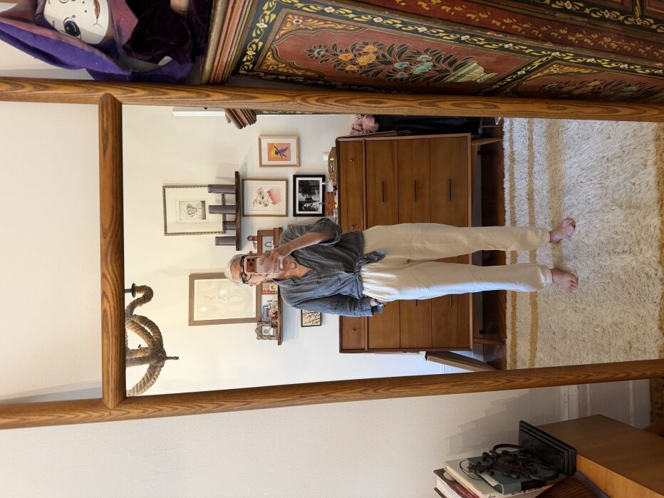Person stands barefoot in front of a mirror, wearing a gray shirt and white pants. A wooden dresser, framed art, and a patterned cabinet appear in the background.