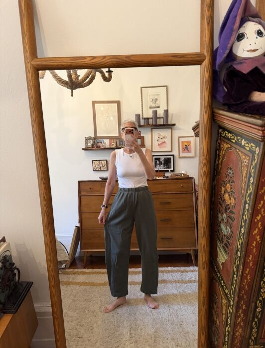 A person stands barefoot in front of a mirror, wearing a white sleeveless top and green pants, taking a selfie in a room with mid-century furniture and framed artwork on the wall.