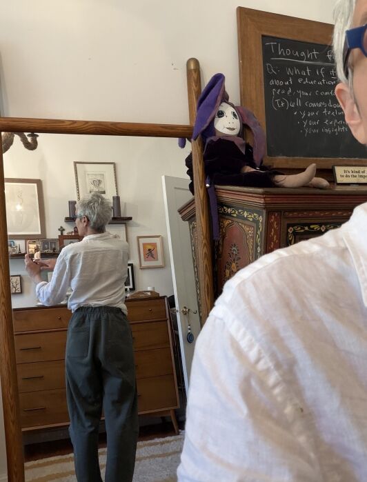 Person with short gray hair and glasses is seen from behind in a mirror, standing in a room with wooden furniture, framed pictures, and a purple-hat doll on top of a cabinet.