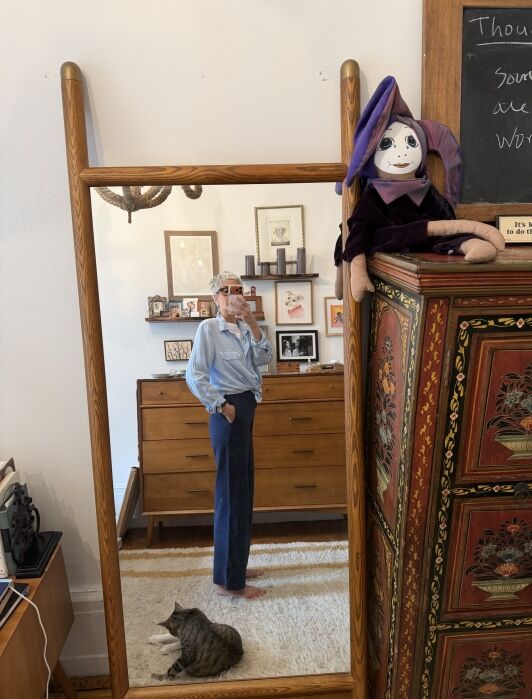 Person taking a mirror selfie in a bedroom with a cat lying on the rug and a doll in a purple outfit sitting on a decorated cabinet.