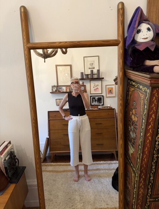 Person takes a mirror selfie in a room with eclectic decor, wearing a black sleeveless top and white pants. Various framed pictures and a large purple puppet are visible in the background.