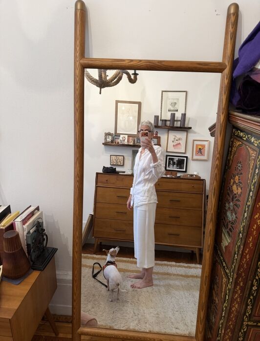 Person in white clothes taking a mirror selfie in a room with a small dog, wooden furniture, art on the wall, and various items on surfaces.