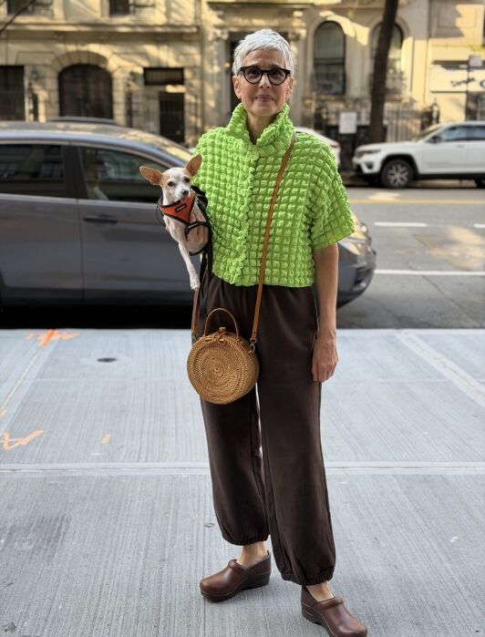 Woman with short gray hair wearing glasses, a bright green textured top, brown pants, and brown clogs stands on a city sidewalk holding a small dog and a round straw bag.