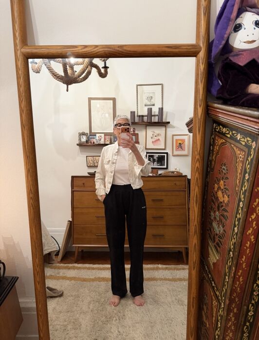 Person stands barefoot in front of a mirror, wearing black pants, a white shirt, and sunglasses. The room has wooden furniture, framed art on the wall, and a decorative cabinet on the right.