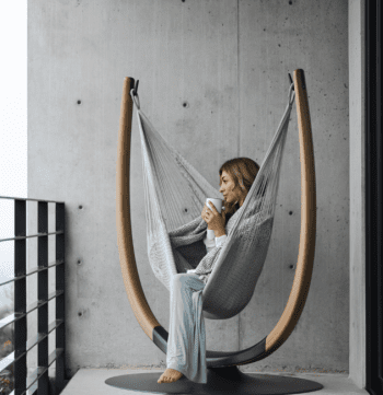 Woman sits in a hammock chair on a balcony, holding a cup and looking out, with a concrete wall and railing in the background.
