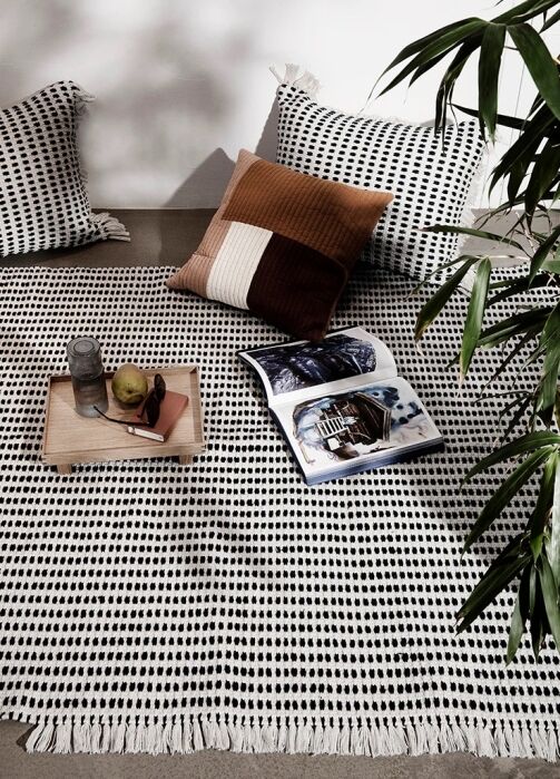 A patterned black and white blanket with two pillows, a tray with fruit and a drink, and an open magazine, set on the floor next to a leafy green plant.
