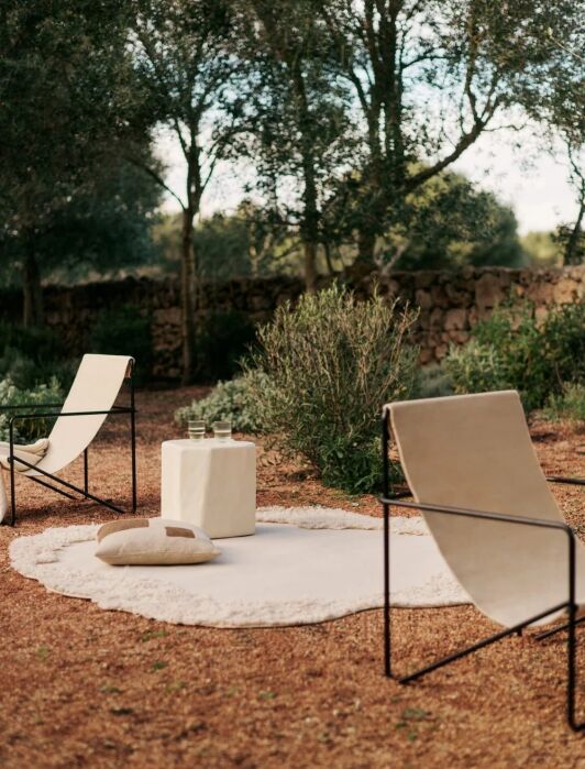 Two canvas lounge chairs and a small white table with glasses are arranged on a round rug outdoors, surrounded by trees and greenery.
