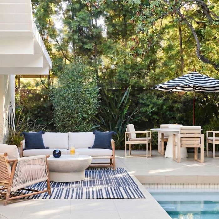 Modern outdoor patio with a seating area, striped rug, wooden furniture, potted plants, and a dining table shaded by a black-and-white striped umbrella next to a pool.