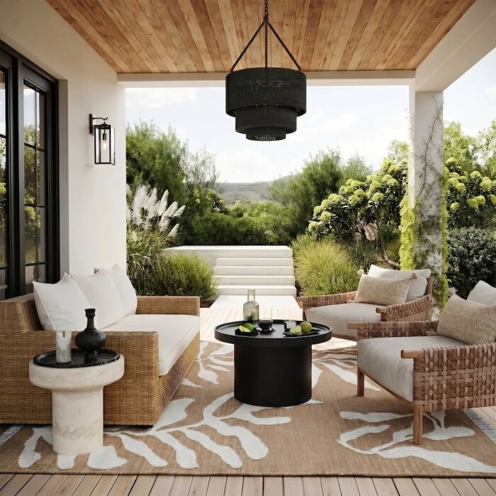 Covered outdoor patio with wicker furniture, black table, neutral cushions, a patterned rug, pendant light, and lush greenery in the background.