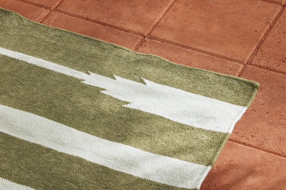 A green and white striped rug with a jagged white pattern on one stripe lies on a reddish-brown tiled floor.