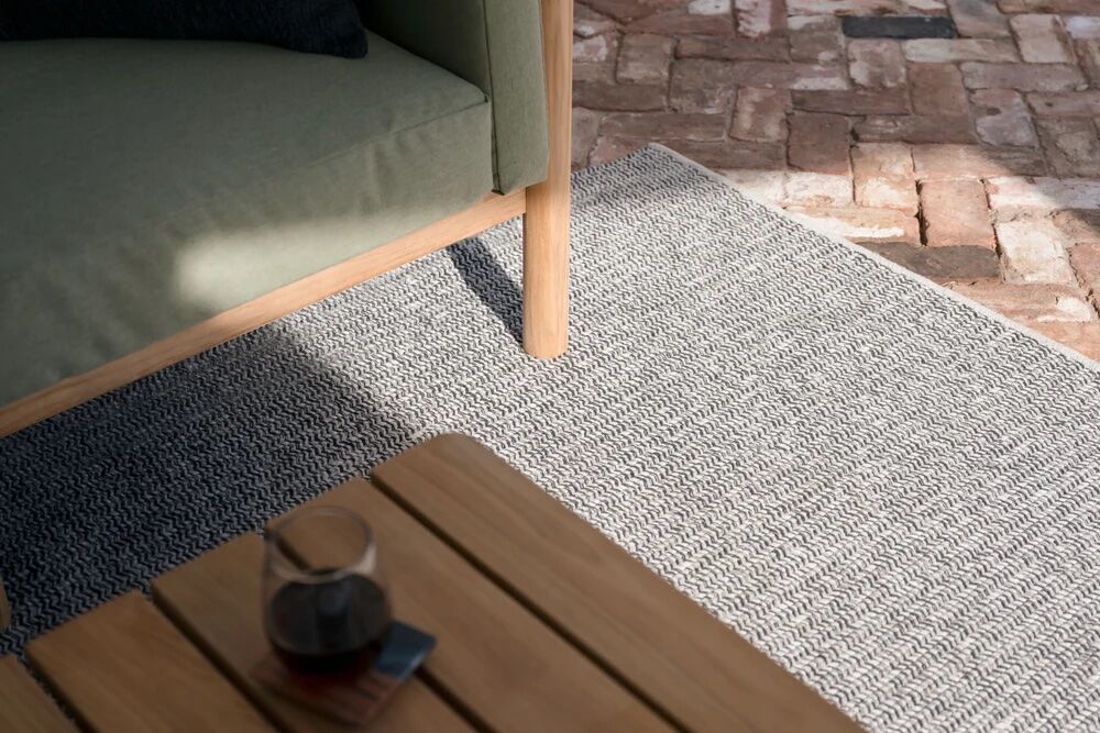 Outdoor seating area with a wooden table, glass of dark liquid, and a book on a textured grey rug, next to a green-cushioned chair on a brick patio.