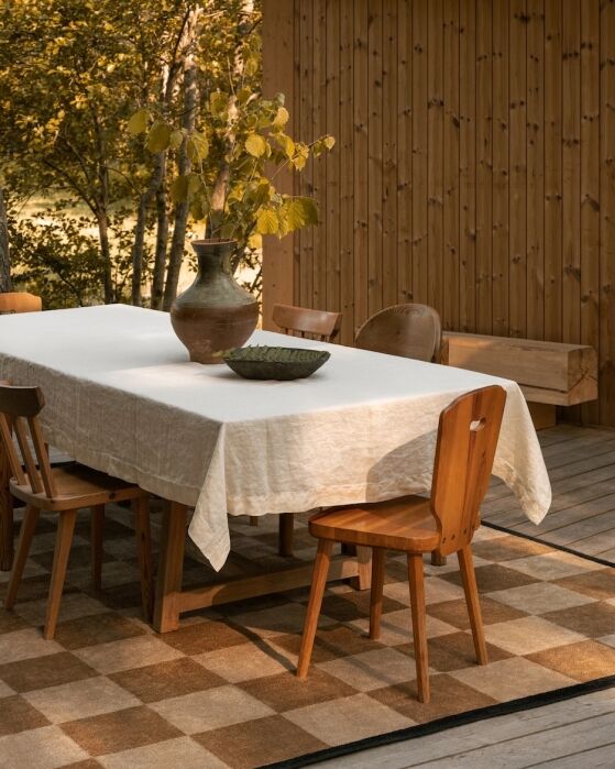 A wooden dining table with a white tablecloth, ceramic vase with branches, and bowl, surrounded by wooden chairs on a checkered rug in a room with wood-paneled walls.
