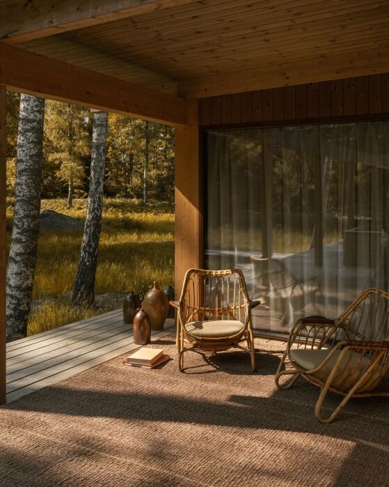 A covered wooden patio with wicker chairs, a small table, vases, books, and glass doors, overlooking a grassy area with trees in warm sunlight.