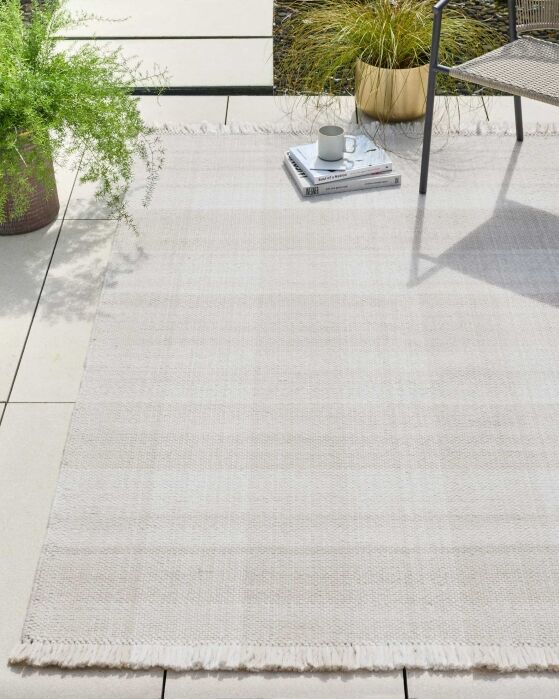 A light-colored woven rug is laid on a tiled patio. There is a chair, two potted plants, and a stack of magazines with a mug on top.