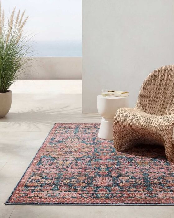 A patterned pink and blue area rug is placed on a light floor beside a woven chair, white side table, and potted grass plant in a minimal, sunlit room.