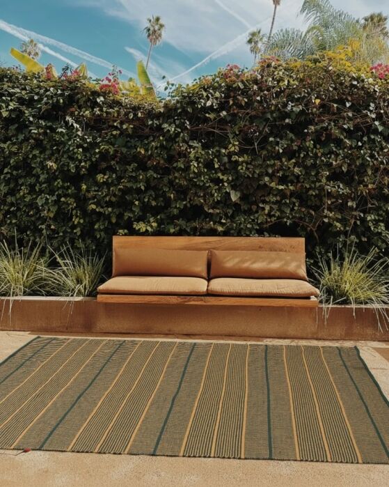 Tan outdoor sofa with two cushions sits in front of a leafy green hedge, with a striped rug and various plants visible under a blue sky.