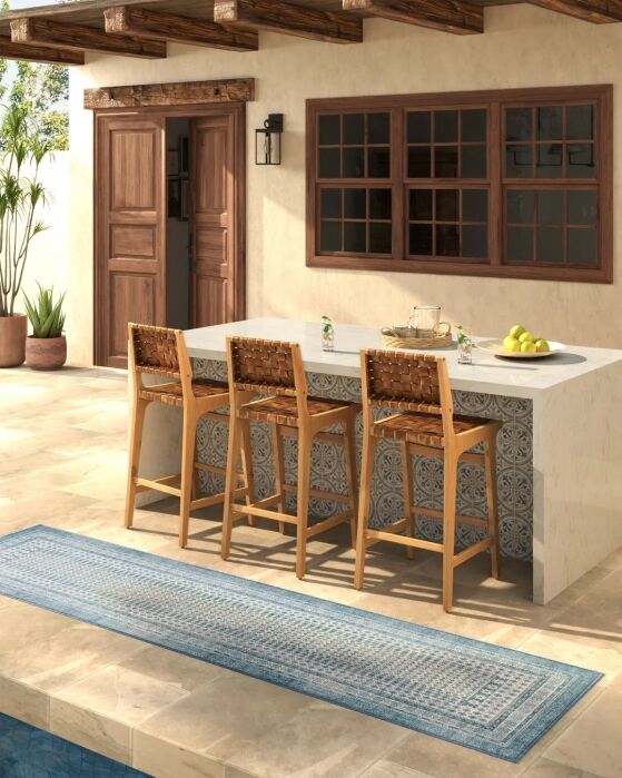 An outdoor patio features a stone-topped bar with three wooden barstools, a patterned rug, and a table set with drinks and fruit, next to a building with wooden doors and windows.