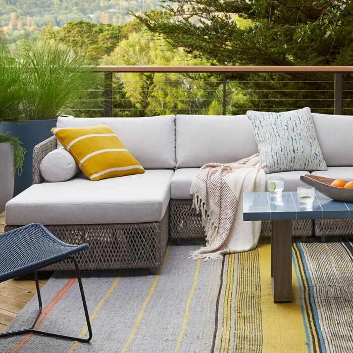Outdoor patio with a gray sectional sofa, two patterned pillows, a throw blanket, a blue table with drinks, a fruit bowl, and a striped rug, surrounded by greenery.