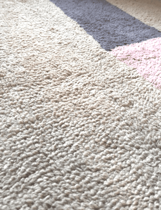 Close-up of a textured beige carpet with sections of gray and pink in the background, illuminated by natural light.