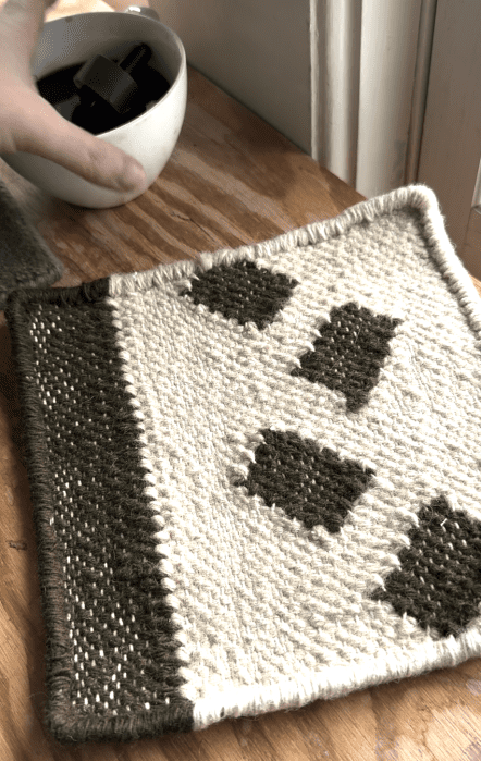 A hand holds a white cup near a woven, rectangular, cream and olive green placemat with geometric patterns, placed on a wooden surface.