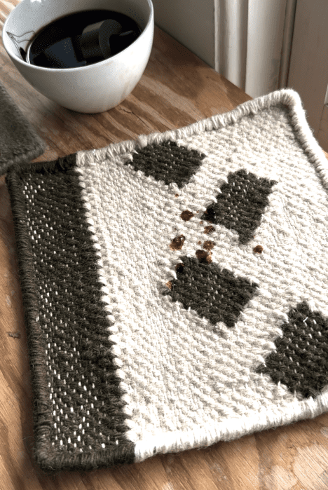 A white and green woven coaster with spilled coffee drops on it sits on a wooden table next to a white cup of black coffee.