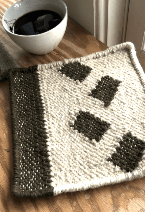A woven coaster with green and cream geometric patterns sits on a wooden surface near a white cup filled with a dark liquid.