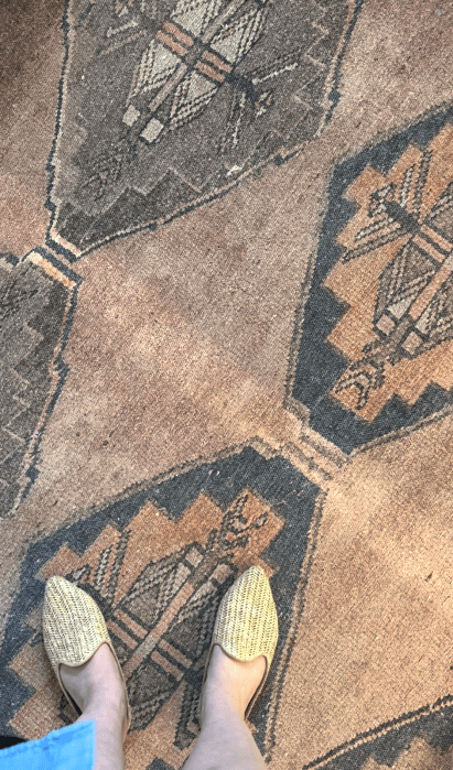 Person wearing woven shoes standing on a patterned rug with geometric designs and earthy tones.