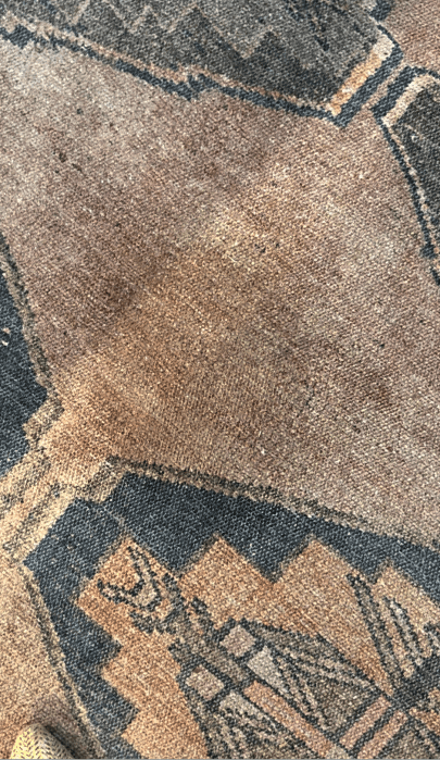 Close-up of a worn, patterned rug with faded earth-tone colors and geometric shapes, showing visible texture and signs of use.