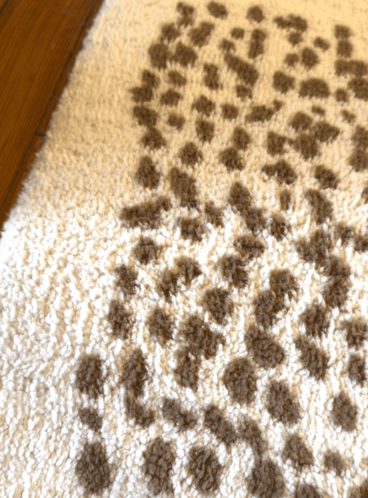 Close-up of a white textured rug with irregular brown spotted patterns, next to a wooden floor.