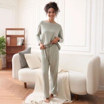 Woman standing barefoot in a living room, wearing light gray loungewear and smiling, in front of a cream sofa with pillows and a blanket, with a wooden shelf and plant in the background.