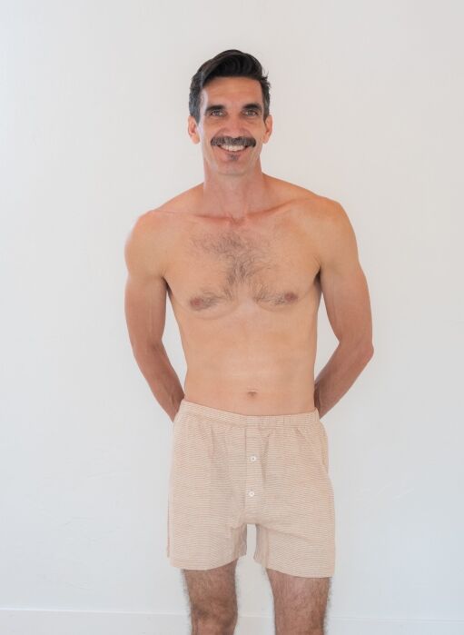 A smiling, shirtless adult man with a mustache stands against a plain white background, wearing light beige boxer shorts, with his hands behind his back.