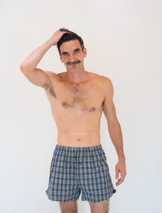 A shirtless man with a mustache wearing plaid boxers stands against a plain white background, raising one hand to his head.