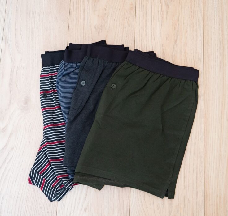 Three pairs of men’s boxer shorts—striped, dark blue, and olive green—are neatly folded on a light wooden floor.