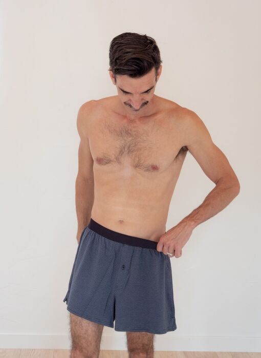 A shirtless man with short dark hair stands indoors, looking down and pulling the waistband of his blue boxer shorts away from his body.