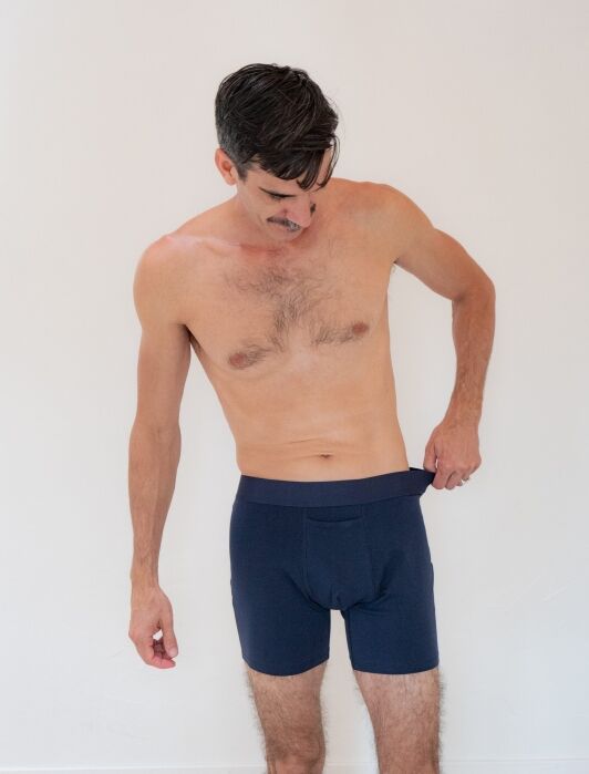 A shirtless man with short, dark hair looks down while adjusting the waistband of his navy blue boxer briefs against a plain white background.