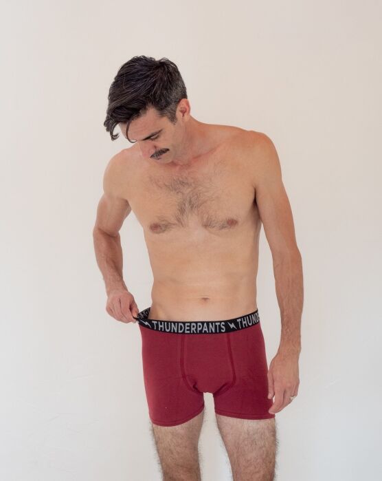 Shirtless man with a mustache looks down while adjusting the waistband of his red Thunderpants underwear against a plain background.