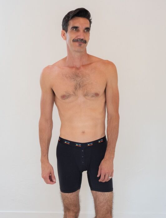 A shirtless man with a mustache stands against a plain white wall, wearing black boxer briefs with a patterned waistband.