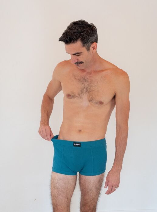 A man with short hair and a mustache stands against a plain background, wearing teal boxer briefs and looking down while pulling the waistband away from his body.