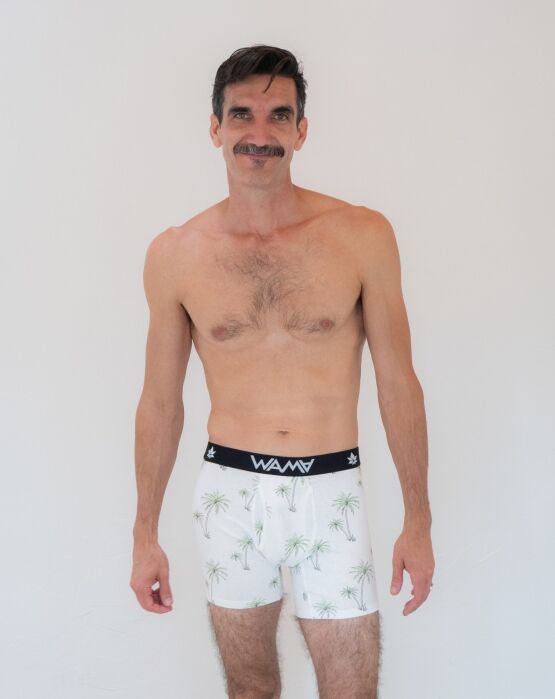 Man with a mustache standing against a plain white background, wearing patterned boxer briefs with a black waistband labeled “WAMA.”.