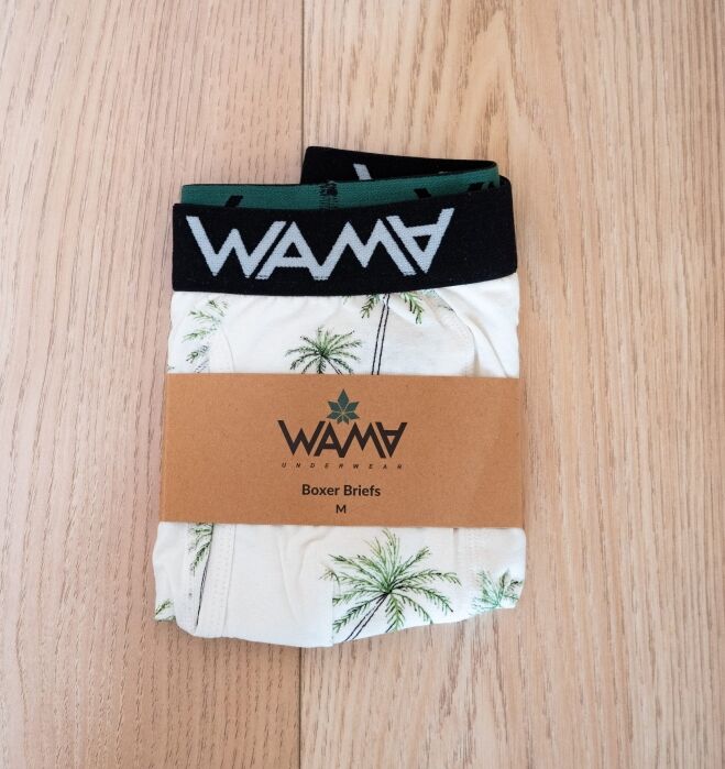 A pair of WAMA brand medium boxer briefs with a palm tree print, folded and wrapped in a brown paper band, displayed on a light wood surface.