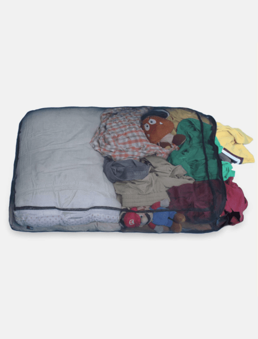 A mesh storage bag contains folded blankets, assorted clothes, and two plush toys, one resembling a mushroom and another resembling a bear.