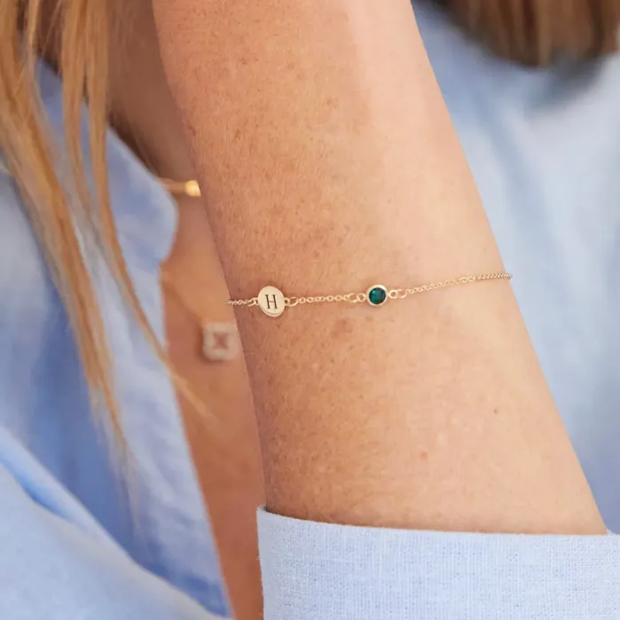 A gold chain bracelet featuring a round charm with the letter H and a small, circular green stone.