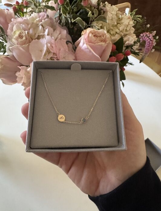 A hand holds a gray gift box containing a delicate gold necklace with a circular charm engraved with the letter “F” and a small blue gemstone.
