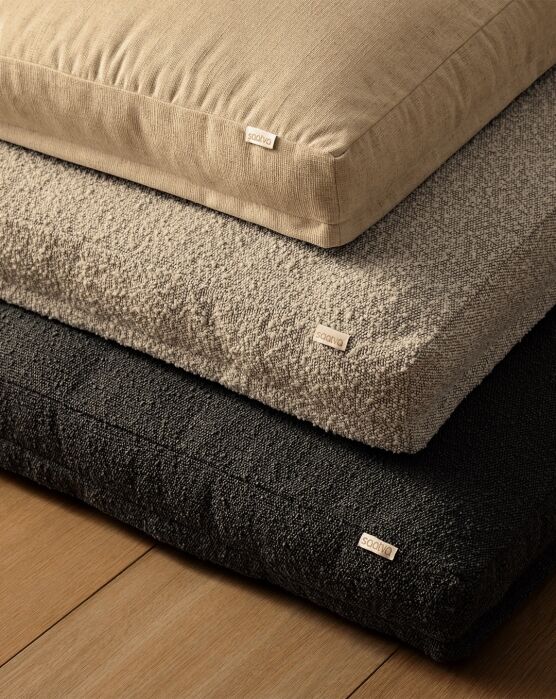 Three rectangular cushions in beige, light gray, and dark gray are stacked on a wooden floor. Each cushion has a small fabric tag with “soavia” on it.