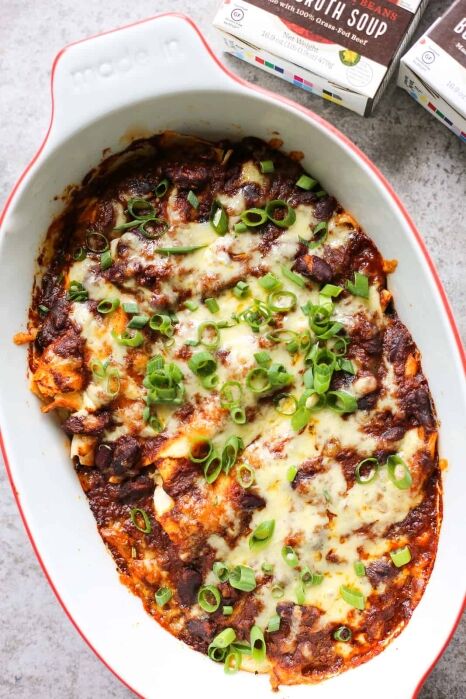 A baked casserole topped with melted cheese, enchilada sauce, and chopped green onions sits in a white oval dish on a light surface.