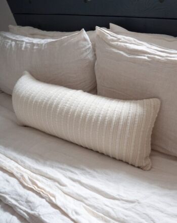 A long, textured white pillow rests on a neatly made bed with two large white pillows and white bedding.